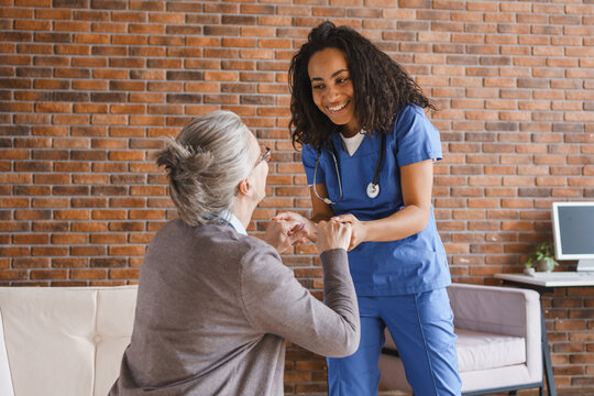 Female caregiver helping elderly woman stand up at home , young african-american nurse holding senior patient by hand, home care, rehabilitation support and mobility assistance concept