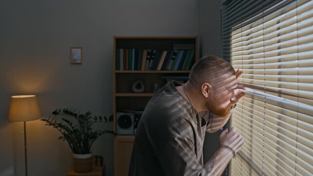 Side view medium shot of Caucasian bearded man peeking through closed window blinds in apartment