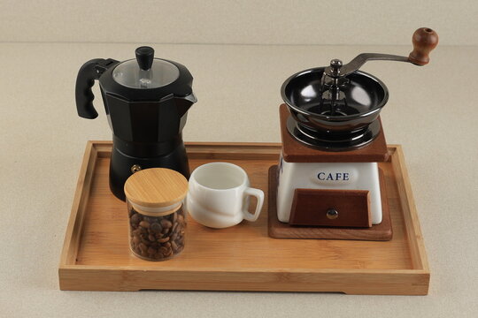 Bird eye view angle of s wooden tray isolated on a clean, minimalist beige background, containing a manual grinder, a mocha pot, a jar of coffee beans, and a small white glass. Bright lighting