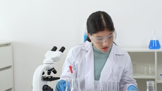 Young Asian women scientist working with test tube in laboratory. Scientist in a lab coat and safety glasses handles a test tube at a desk in a laboratory, make note on laptop, experimentation in a co