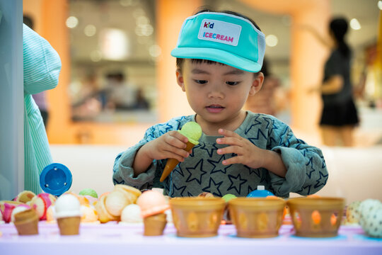 Cute little Asian boy playing role-play as ice cream seller at indoor playground, focused on scooping toy ice cream cone with authentic curious expression.