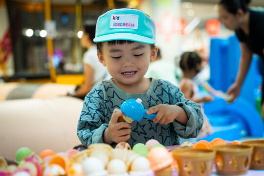 Cute little Asian boy playing role-play as ice cream seller at indoor playground, focused on scooping toy ice cream cone with authentic curious expression.