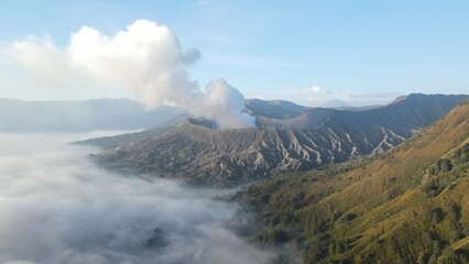 Obraz premium Stunning aerial view of Mount Bromo volcano erupting with white smoke above a thick morning mist.
