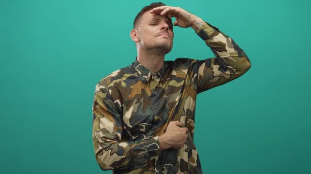 Young man in camouflage shirt, soldier posing with hand on chin and shielding forehead against bright light in studio; duty contemplation.
