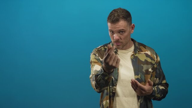Man in camouflage jacket identified as soldier making a beckoning hand gesture with fingers toward camera and touching his chest in a studio; defiance.