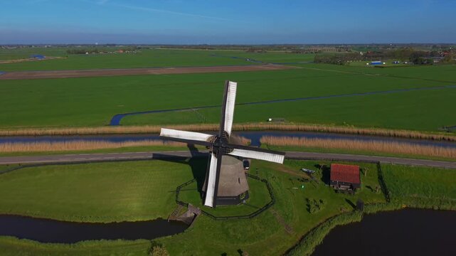 Aerial view of Bovenmolen G windmill standing over green polder fields and a canal under a clear blue sky in Schermerhorn, Noord-Holland, Netherlands.