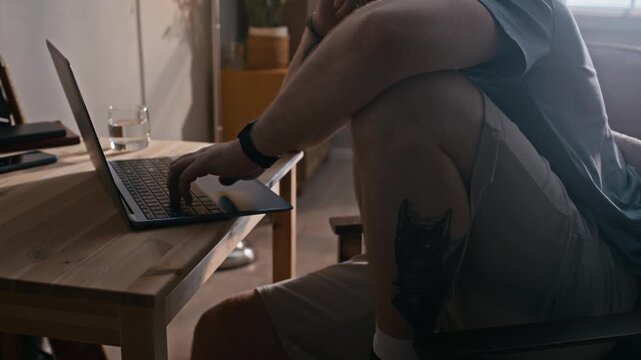 Tilt up of man wearing electronic ankle bracelet working on laptop from home while sitting in cozy apartment during home confinement
