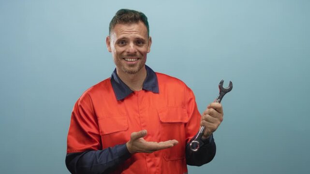 Man handyman young hispanic in orange uniform holding a wrench and gesturing with open palm in studio setting; uncertain assistance.