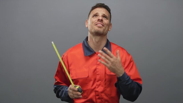 Man in handyman uniform holds a tape measure and displays palms up shrug gesture in studio; uncertainty.