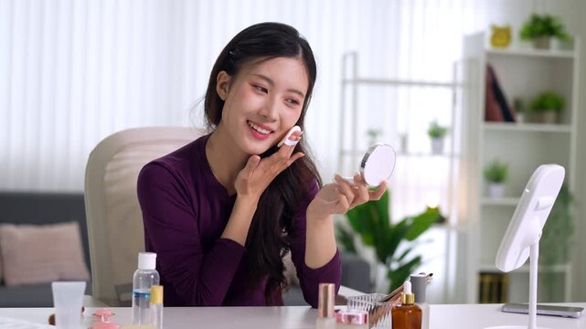 Asian women beauty influencer applying makeup cosmetic product holding mirror recording video tutorial at home workspace creating online content for social