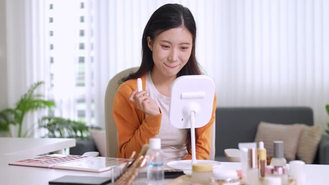 young Asian women beauty influencer applying lipstick makeup tutorial using mirror recording video for social media content creation at home living room with