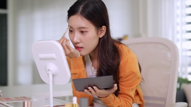 young Asian woman apply makeup with mirror brush eyeshadow palette cosmetic beauty routine morning preparation at home table casual orange sweater selfcare