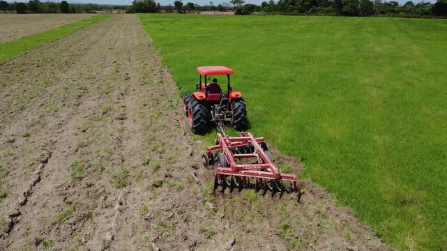 Aerial view of tractor with disc harrow tilling soil beside green rice field in Colombia