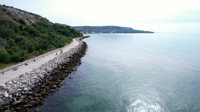 Aerial view of people walking along a stone-reinforced coastal promenade with lush green hills and the calm Black Sea in Balchik, Dobrich Province, Bulgaria.