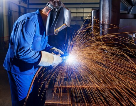 professional welder in protective mask and blue uniform working with metal arc welding machine in industrial factory workshop with bright sparks