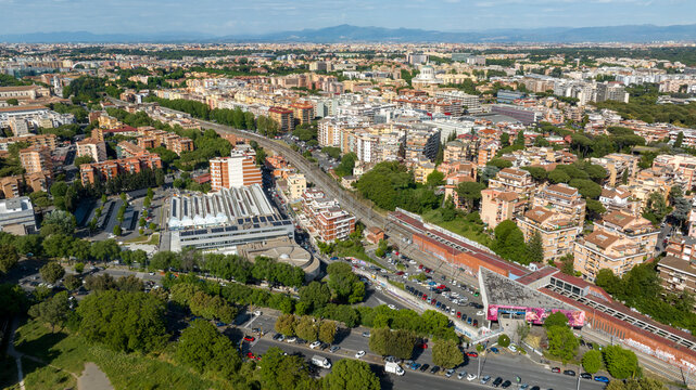Aerial view of a modern urban neighborhood with residential buildings, a railway station, lush green trees, and a city skyline under a blue sky during a sunny day. It's Marconi district in Rome, Italy
