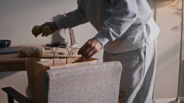 Tilt up of woman with braided hair standing by small table and unpacking grocery products in apartment while wearing electronic ankle bracelet