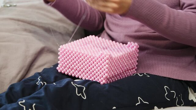 Hands crafting pink beaded box with fine needlework on dark patterned fabric lap