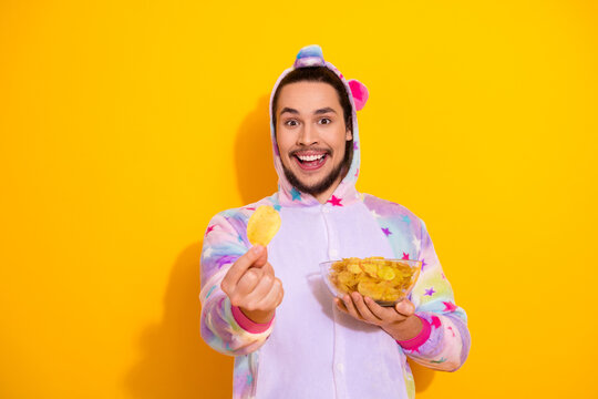 Happy man in colorful unicorn pajama outfit enjoying chips in a bright yellow background