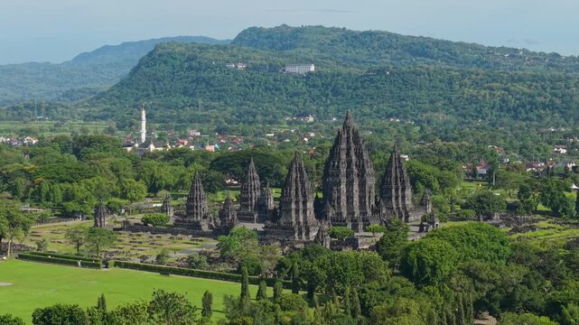 Aerial view of the ancient Hindu Prambanan Temple complex featuring towering stone spires set against a backdrop of forested hills in Kecamatan Prambanan, Daerah Istimewa Yogya, Indonesia.