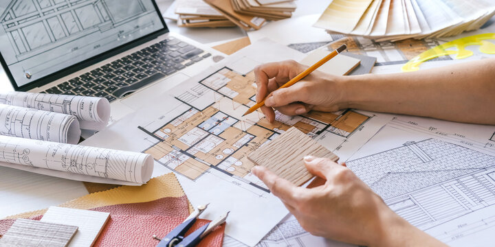 Interior design process with hands selecting wood sample over architectural plans, floor plan, laptop and color palette, residential renovation planning and material selection concept.