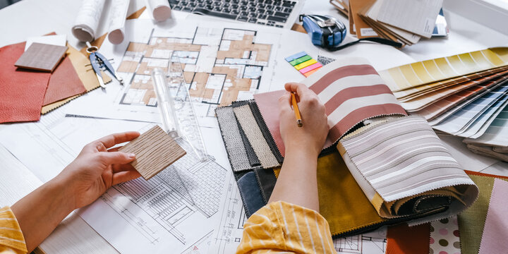 Interior design process with hands selecting wood sample over architectural plans, floor plan, laptop and color palette, residential renovation planning and material selection concept.
