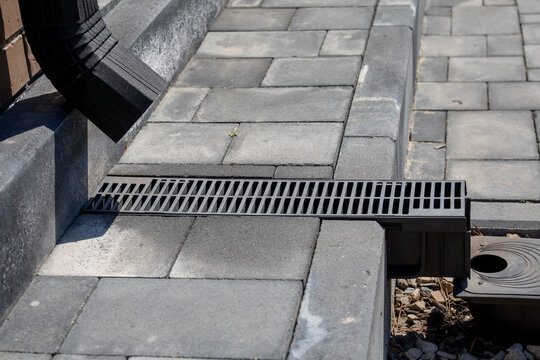 Linear drainage grate installed between concrete pavers, connected to downspout and catch basin, residential hardscape water management system, spring maintenance