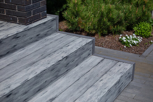 Gray composite deck stairs next to dark brick column, mulched garden bed with pine shrub and white flowers, modern residential outdoor hardscape design