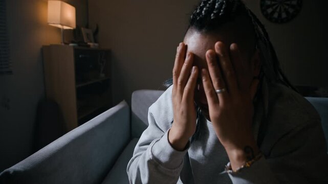Dolly in portrait of Biracial woman wearing ankle monitor sitting on sofa with hands covering her face before looking at camera, expressing stress, emotional struggle and vulnerability in home setting