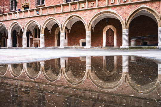Courtyard of Collegium Maius in Krakow with late Gothic arcades and cloisters on a cloudy day after rain, with architectural reflections visible in puddles