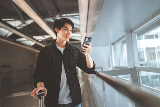 Joyful young Asian male traveler using a smartphone to check flight or book a ride at a modern airport skywalk. Carrying a suitcase and backpack, he enjoys seamless travel. Perfect for digital nomad, 