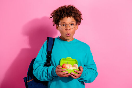 Surprised school student holding a green lunchbox with apple and snacks while wearing backpack against pink background