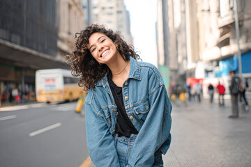 Obraz premium A young woman in a denim jacket smiles brightly on a city street, surrounded by tall buildings and blurred urban activity.