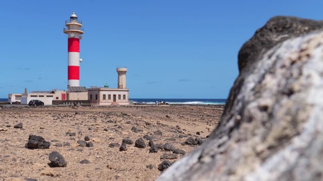 El Toston lighthouse near El Cotillo with lava coast and large foreground rock, Atlantic Ocean and blue sky in background.