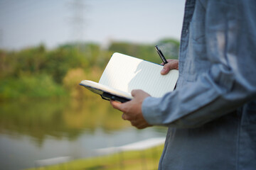 Young man sitting on the grass writing in a notebook and lying down with a book over his face,...