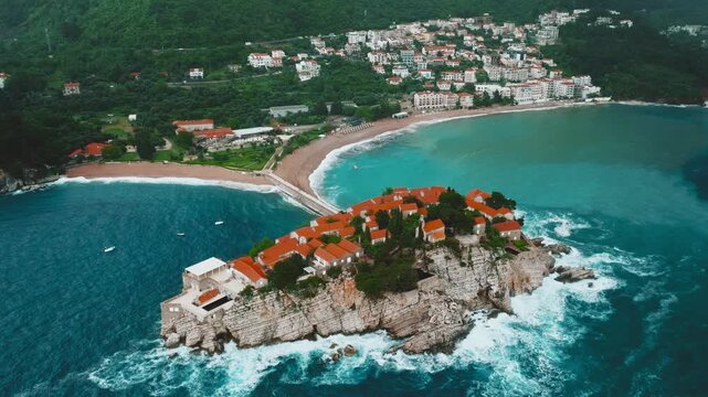 Cinematic aerial drone footage of Sveti Stefan featuring the iconic island village connected to the mainland by a narrow causeway, surrounded by turquoise Adriatic waters