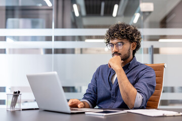 A thoughtful young Indian man is sitting at a desk in the office and looking at the laptop screen