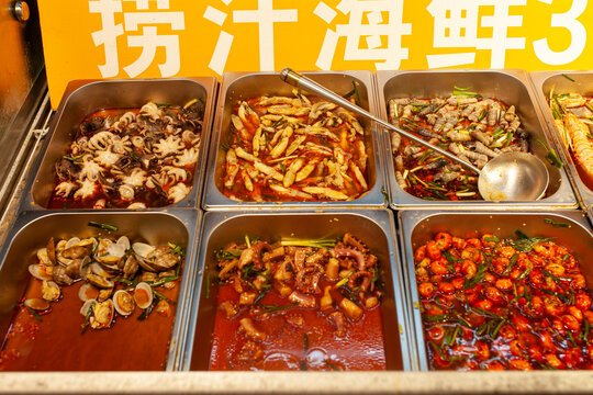 Close-up photo of six stainless-steel trays filled with spicy Asian seafood dishes: octopus, clams, squid and assorted shellfish in red sauces. Street-food culture and market dining experiences.