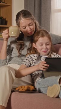 Vertical shot of adult woman drinking coffee while watching film on tablet with preteen daughter, both lying back on couch in living room