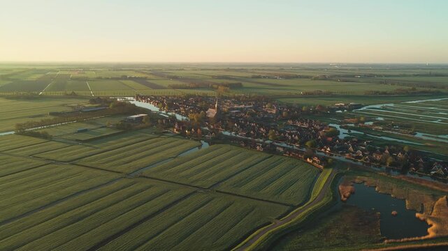 Aerial view of the historic village of Schermerhorn with its church spire surrounded by green polder fields and canals during golden hour in Schermerhorn, North Holland, Netherlands.