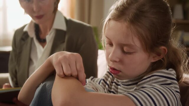 Close up shot of upset preteen girl embracing knees while struggling with emotions in psychotherapy session then female therapist comforting child with supportive shoulder touch during home visit