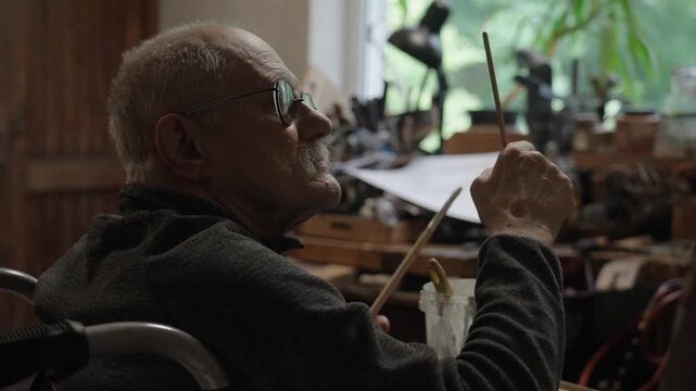 Senior artist on a wheelchair paints an oil painting on canvas in a studio