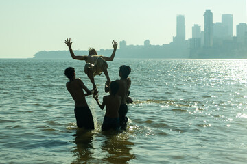 Fototapeta premium Silhouettes of young Indian men playing in ocean at the beach. Group of people throwing their friend into the air while playing in the water during sunset