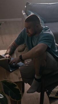 Vertical eye level shot of man under home confinement working on laptop at small desk while wearing electronic ankle tracker