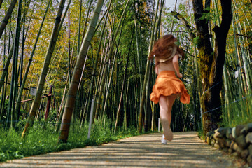 Fototapeta premium Woman running on a sunlit bamboo forest path in an orange skirt, a back view capturing motion and joyful movement along a leafy trail surrounded by tall green trees.