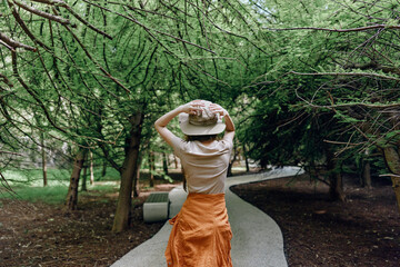 Naklejka premium Woman walking on a winding path through a forest under thick trees, wearing a hat and orange skirt, back view in nature with green canopy, peaceful outdoor exploration scene.