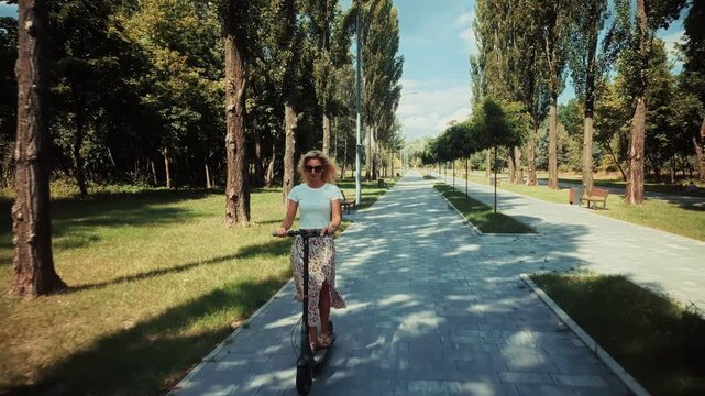 Woman Riding Electric Scooter On Sunny Alley In City Park. Eco Friendly Transportation And Modern Urban Mobility