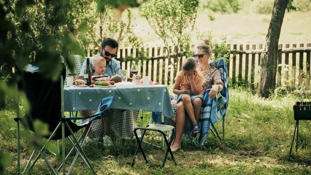 Happy Family Having Picnic Dinner In Garden.  Parents With Children During Summer Barbecue Party. Joyful Moments Of Togetherness