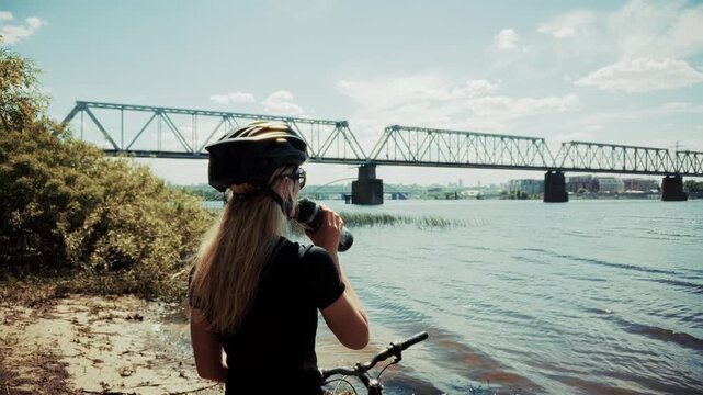 Cyclist Drinking Water from Bottle while Standing with Bicycle on Riverbank. Workout and Hydration during Bicycling Trip