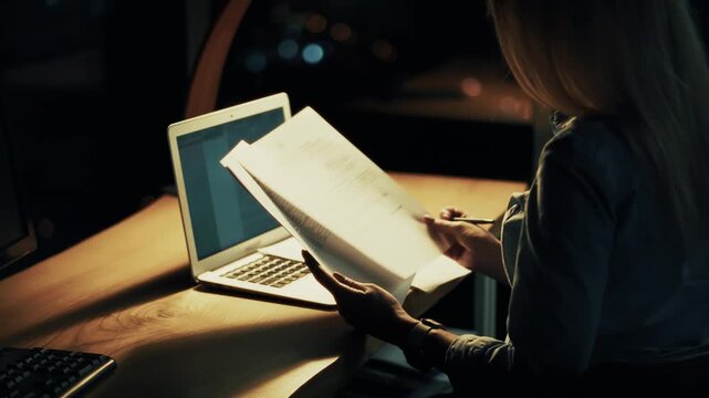 Working Overtime In Office At Night. Secretary Reading Paperwork Near Laptop Under Lamp